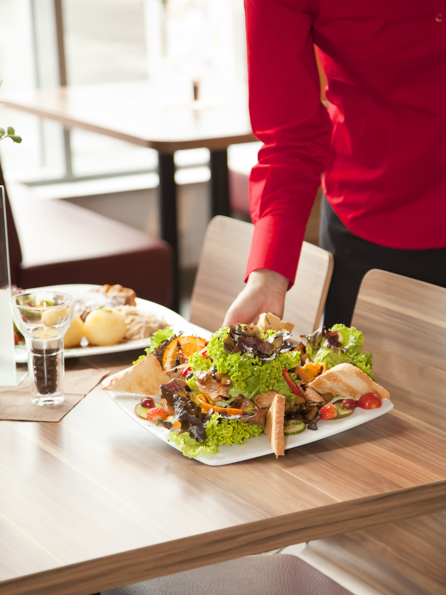 Servicepersonal mit roter Bluse serviert geschmackvollen Salat mit Putenstreifen, dazu geröstetes Brot. Im Hintergrund Braten mit Knödel