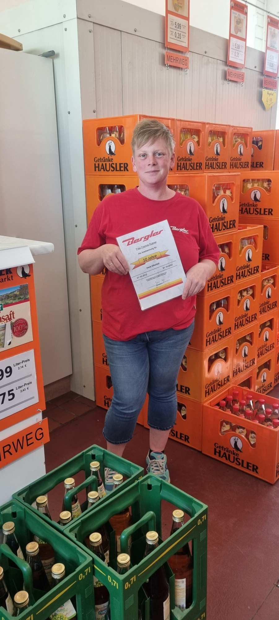 Im Vordergrund die Jubilarin Frau Rubner mit rotem T-Shirt und ihrer Urkunde. Sie befindet sich im angrenzenden Getränkeshop der Bergler Tankstelle in Erbendorf. Im Hintergrund orange Getränkekästen.