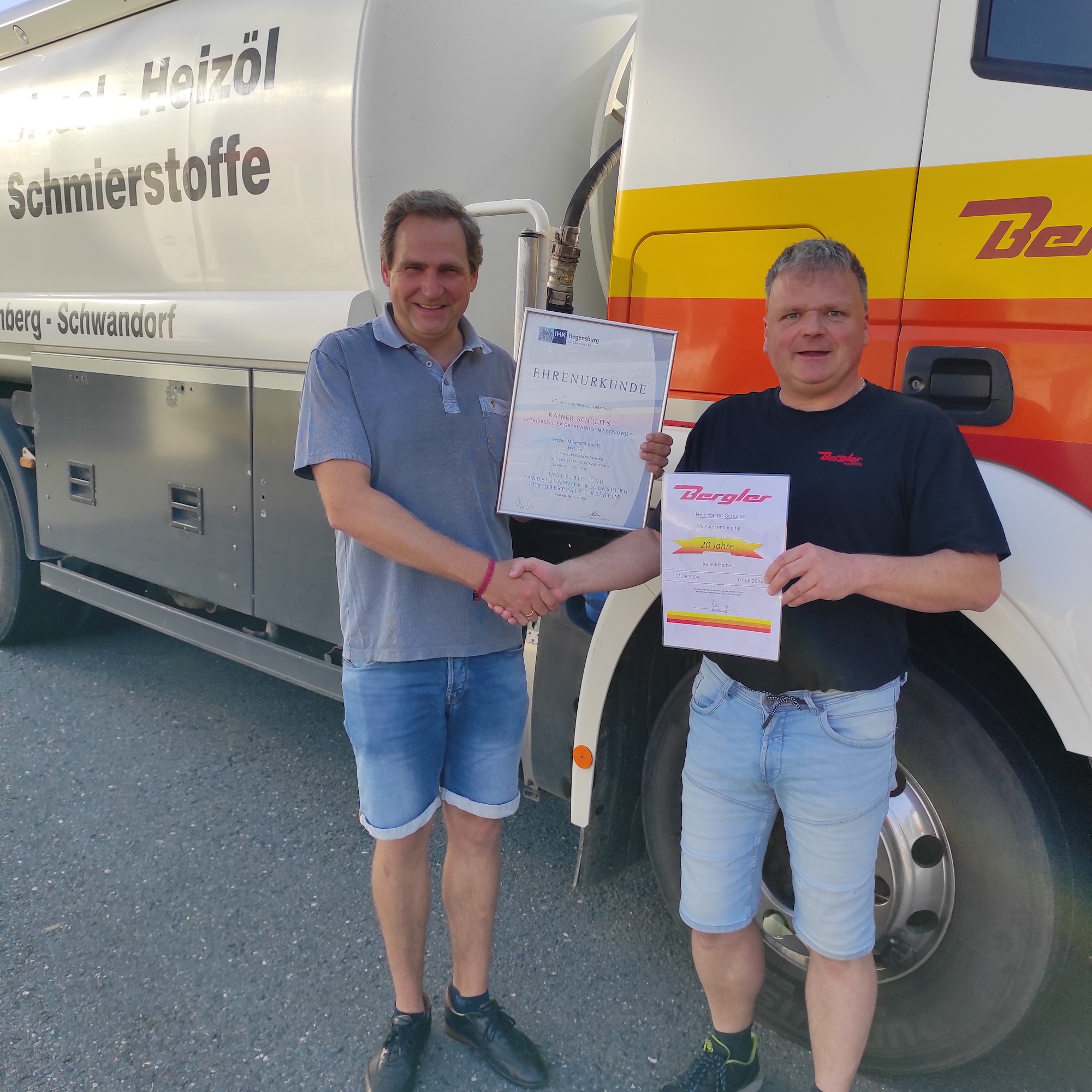 Geschäftsführer Gerhard Bergler, links, übergibt Rainer Schultes, der rechts neben ihm steht, die Urkunde. Im Hintergrund ein Sattelzug der Firma Bergler.