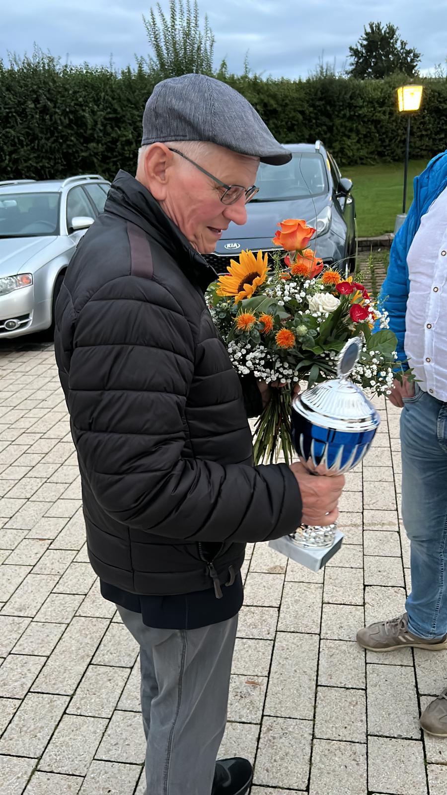 Älterer Mitarbeiter mit Mütze hält Blumenstrauß und Ehrenpokal auf dem Firmenparkplatz