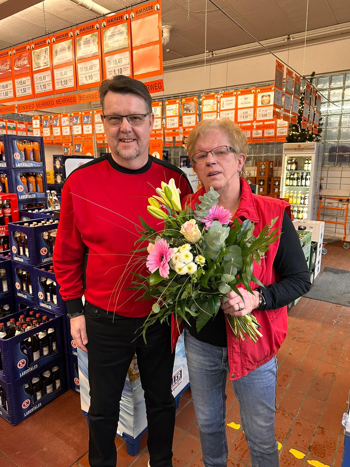 Peter Schneider, links im Bild, und Heidi Mittelmeier stehen vor dem Getränkemarkt der Station in Erbendorf. Beide haben die rote Bergler-Kleidung an, Frau Mittelmeier hat einen großen Blumenstrauß in der Hand