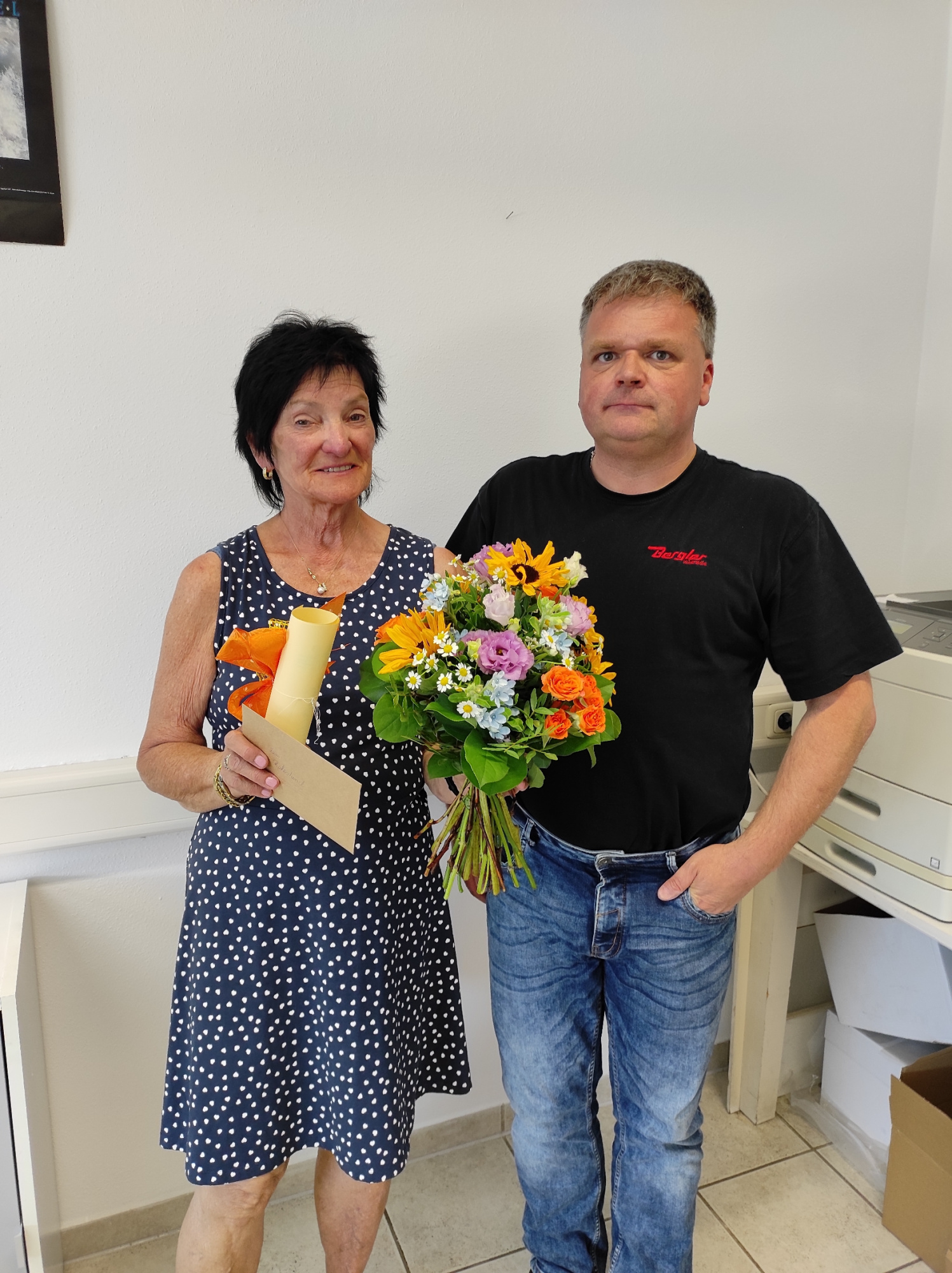 Links Fr. Ackermann mit einem Kuvert und Blumenstrauß in der Hand, das Präsent wurde von Hr. Schultes überreicht, der rechts neben ihr im Bild steht. Beide stehen vor einer weißen Wand und lächeln in die Kamera.