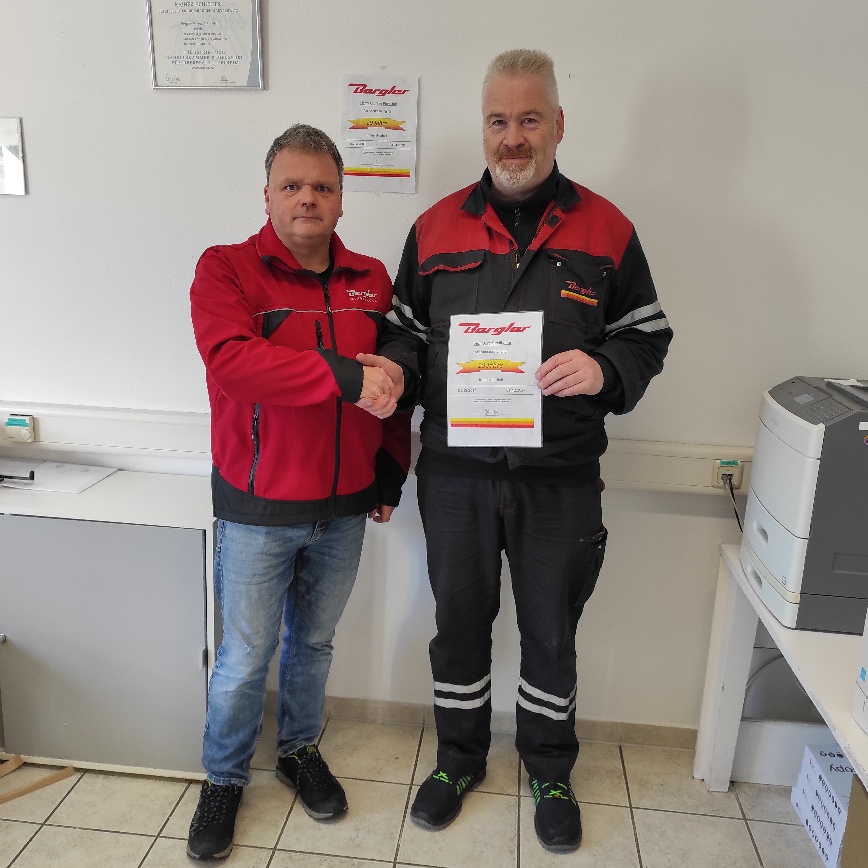 Rainer Schultes in roter Bergler-Jacke und blauer Jeans schüttelt Jörg Stadlbauer in schwarz-roten Arbeitsklamotten die Hand und gratuliert ihm zum Jubiläum. 